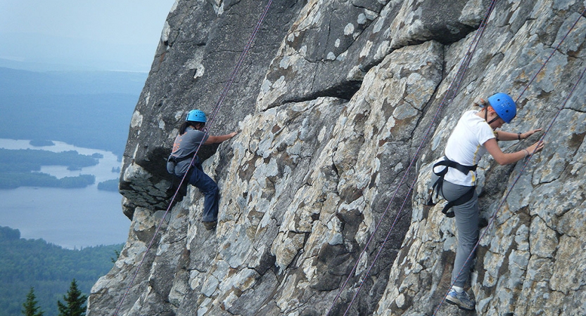 Two people wearing safety gear are secured by ropes as they each climb a rock face. 